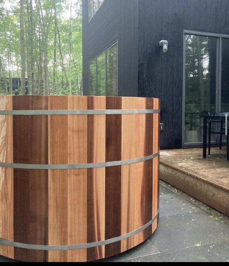 Classic round cedar hot tub beside a modern black house in the woods